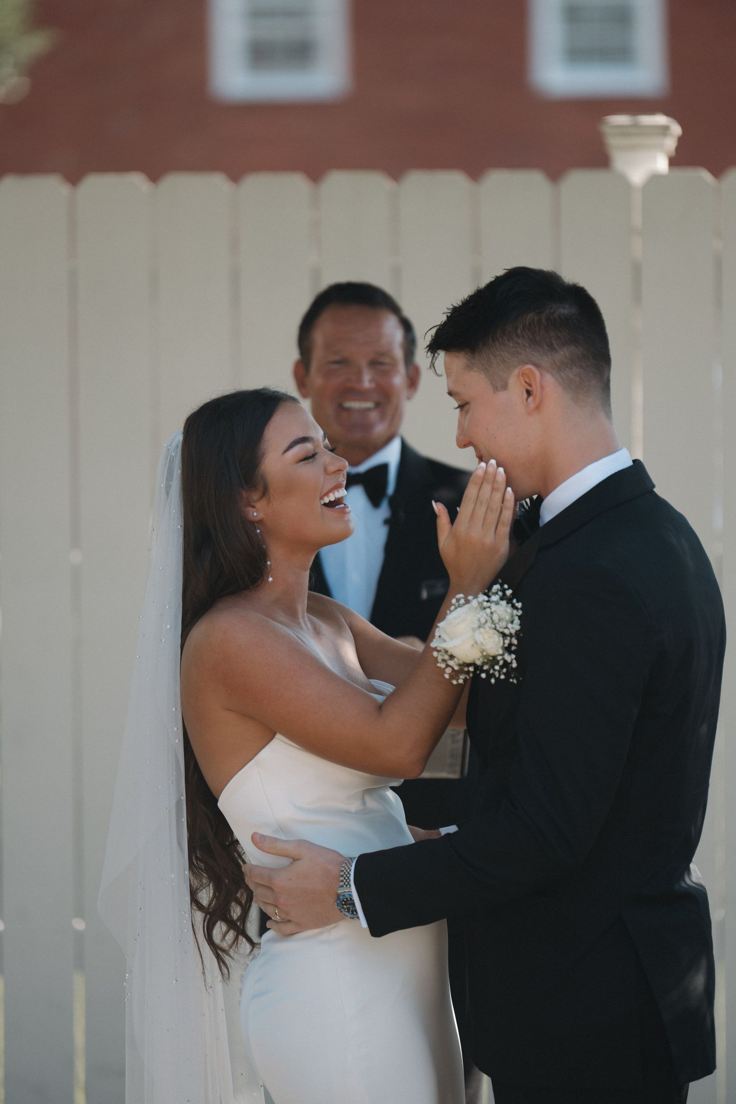 Laughing bride in sleek white gown touches grooms face during outdoor wedding ceremony with officiant behind them.