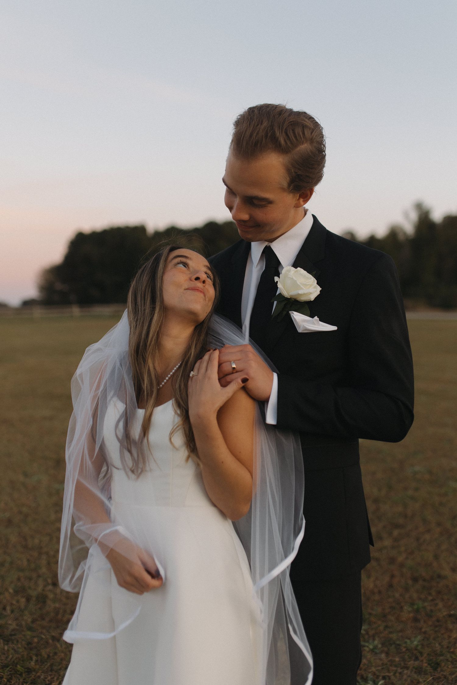 Groom in black suit embraces bride in white dress and veil outdoors at golden hour.
