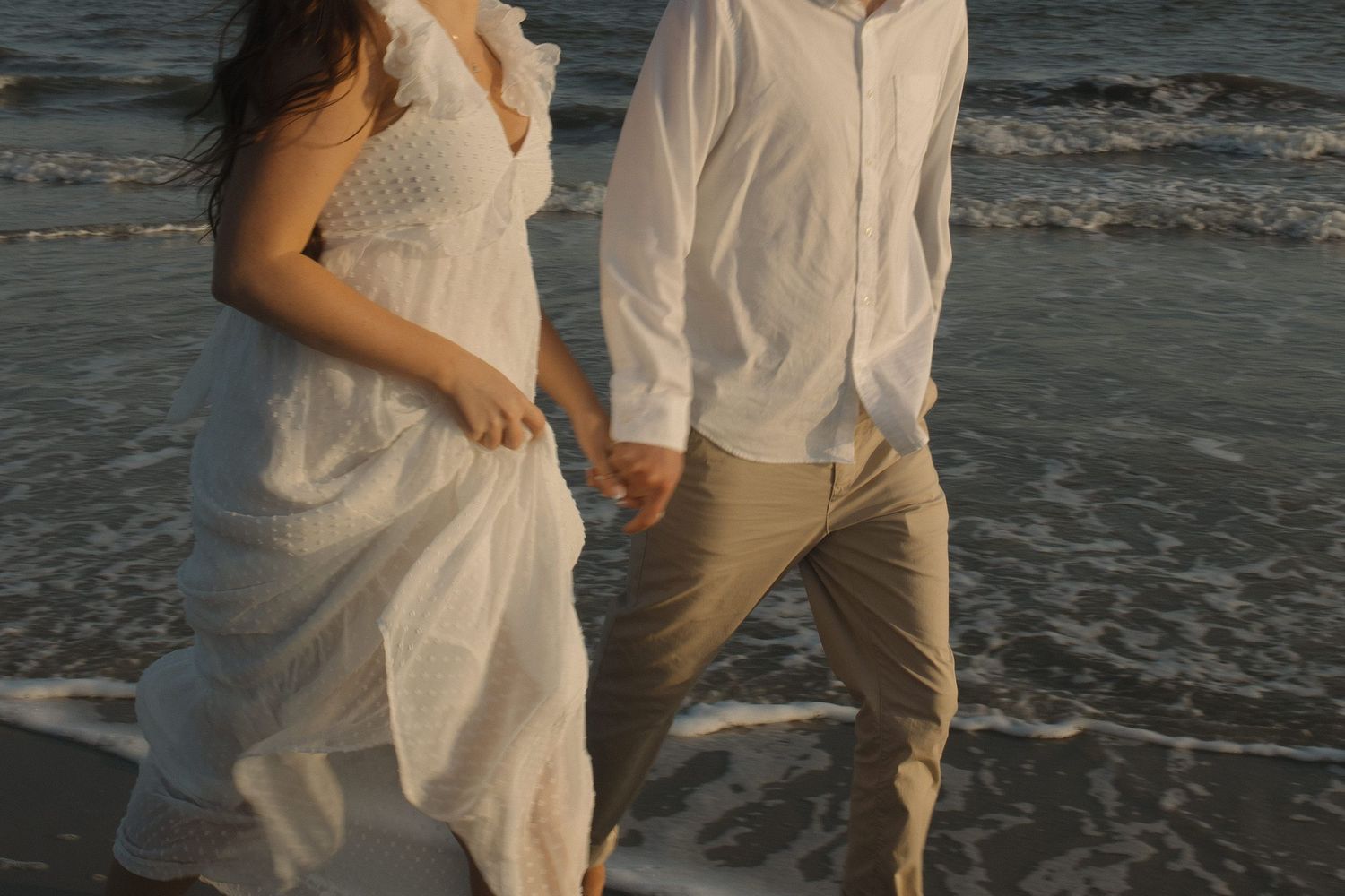 Bride and groom walk hand in hand along ocean shoreline during golden hour beach wedding shoot.