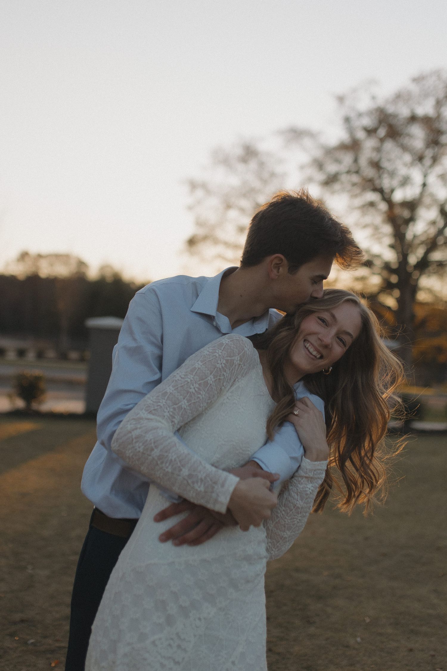 Man embracing woman in lace dress from behind, both laughing joyfully at sunset outdoors.
