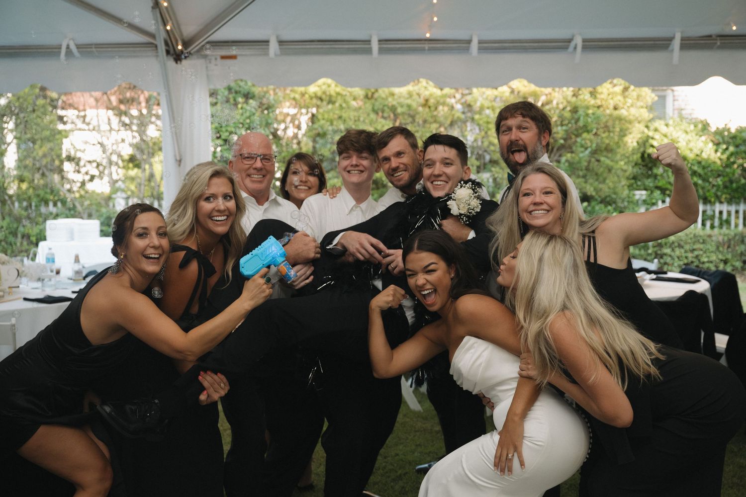 Wedding party group selfie with bride in white dress and guests in black attire celebrating under outdoor tent.