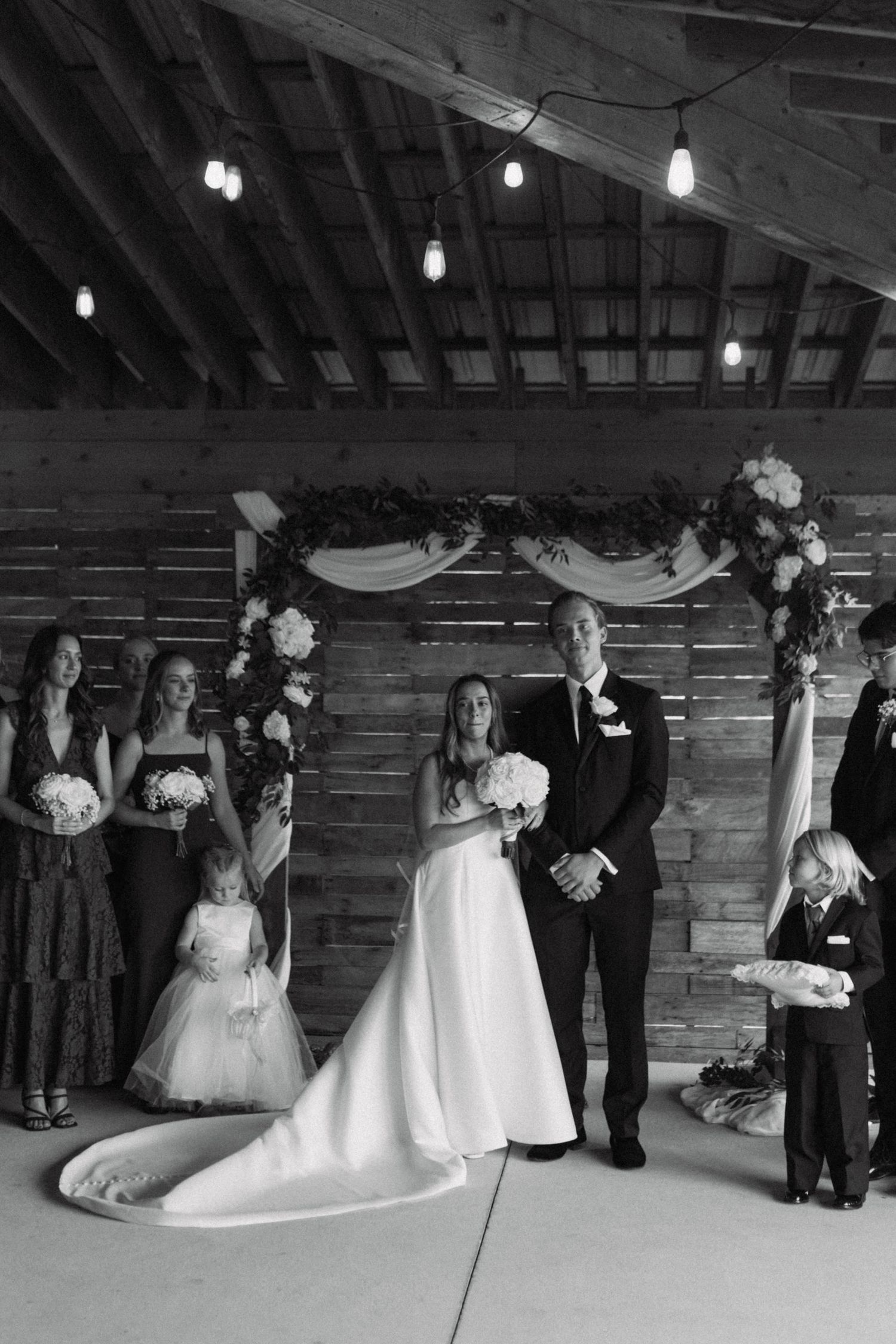 Black and white photo of bride and groom at rustic barn wedding ceremony with bridesmaids and flower girl under floral arch.
