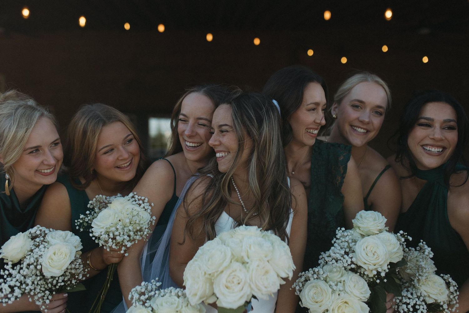Bride and bridesmaids smiling together holding white rose and baby's breath bouquets at an outdoor wedding celebration.
