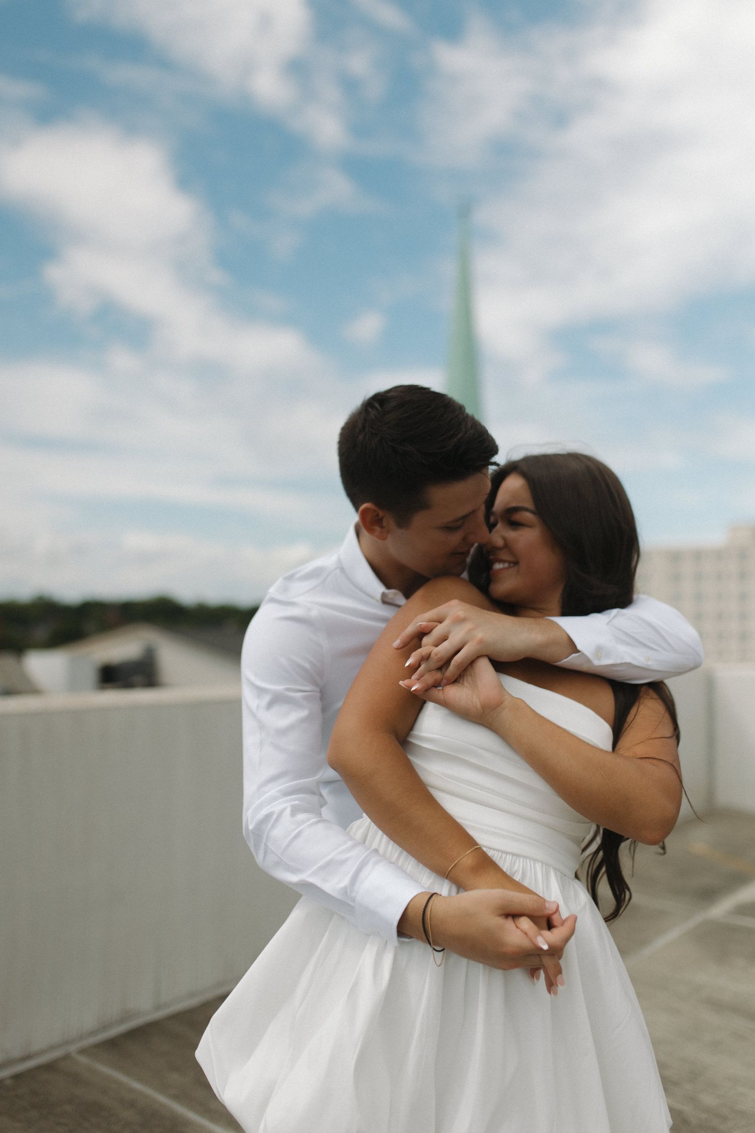 Happy couple in white outfits hugging and laughing on a rooftop with a church steeple behind.