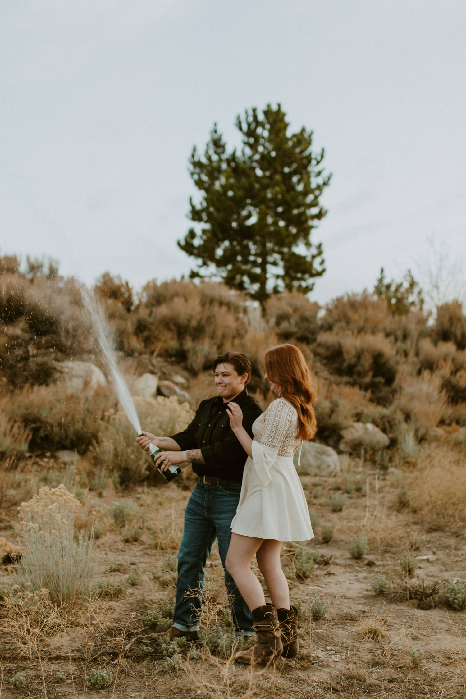 Couple celebrating with champagne spray in rustic outdoor setting with pine trees.