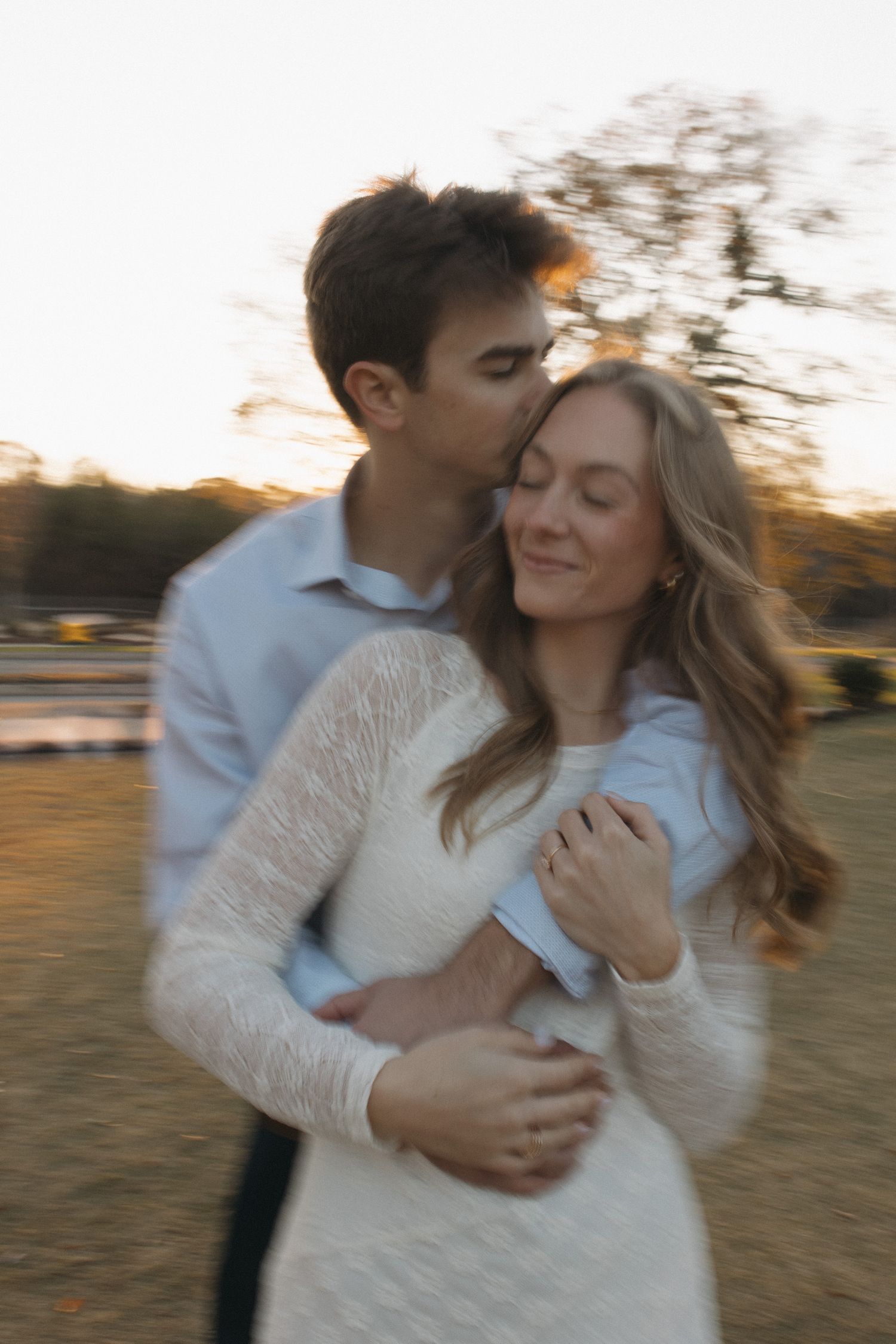 Couple shares a tender embrace at golden hour, man kissing womans temple in a blurred bokeh setting.