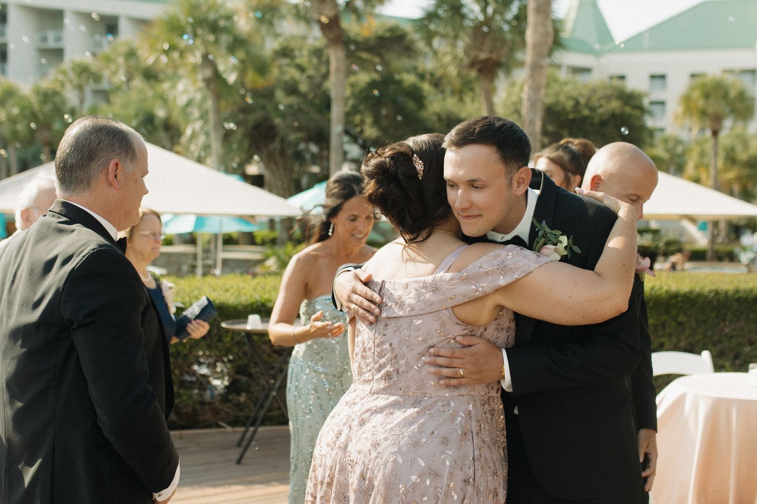 Groom in tuxedo embracing wedding guest in sparkly dress at outdoor wedding reception with tropical backdrop.