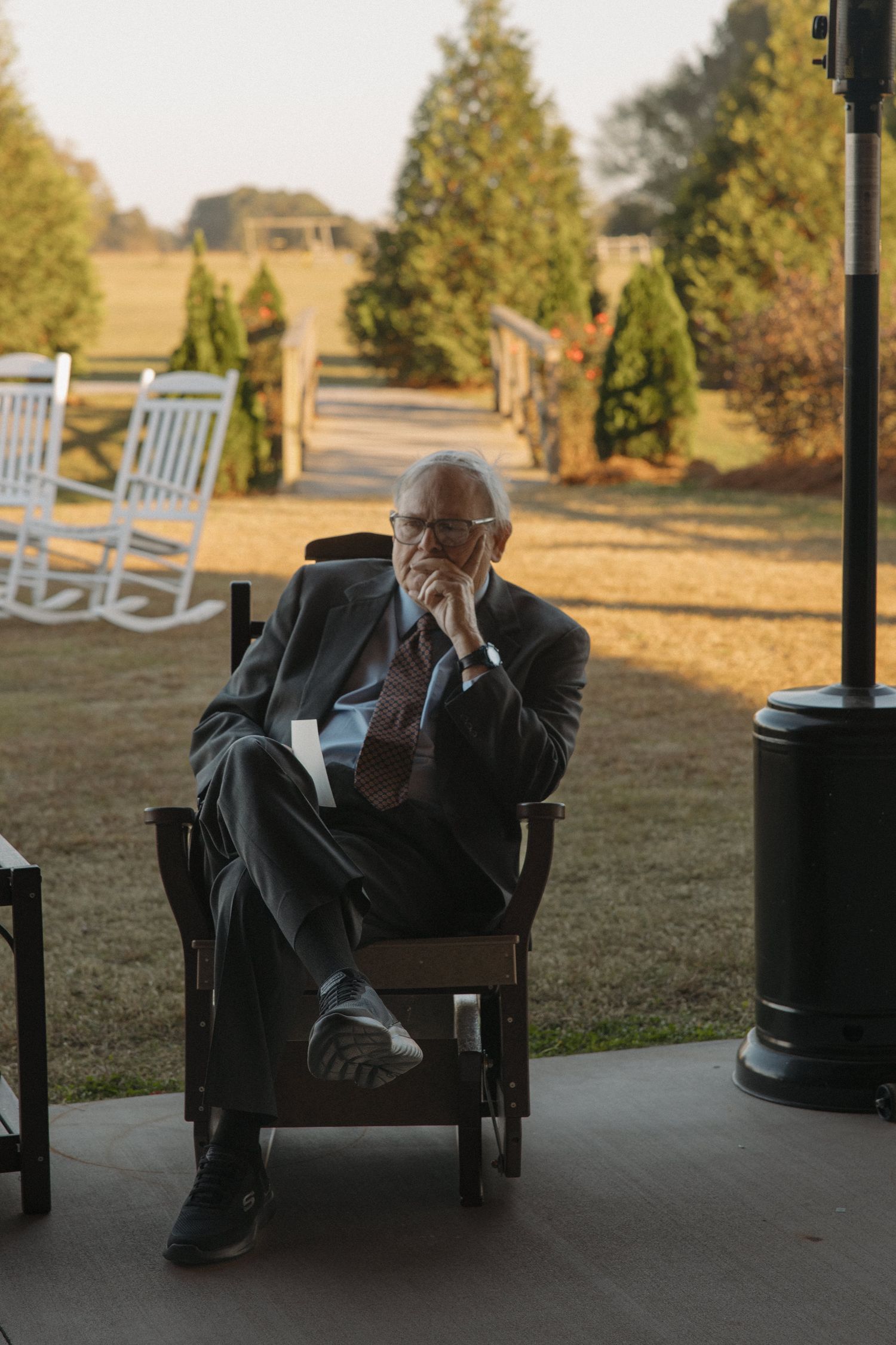 Elderly man in suit sitting thoughtfully in wooden chair on porch overlooking green lawn at sunset.