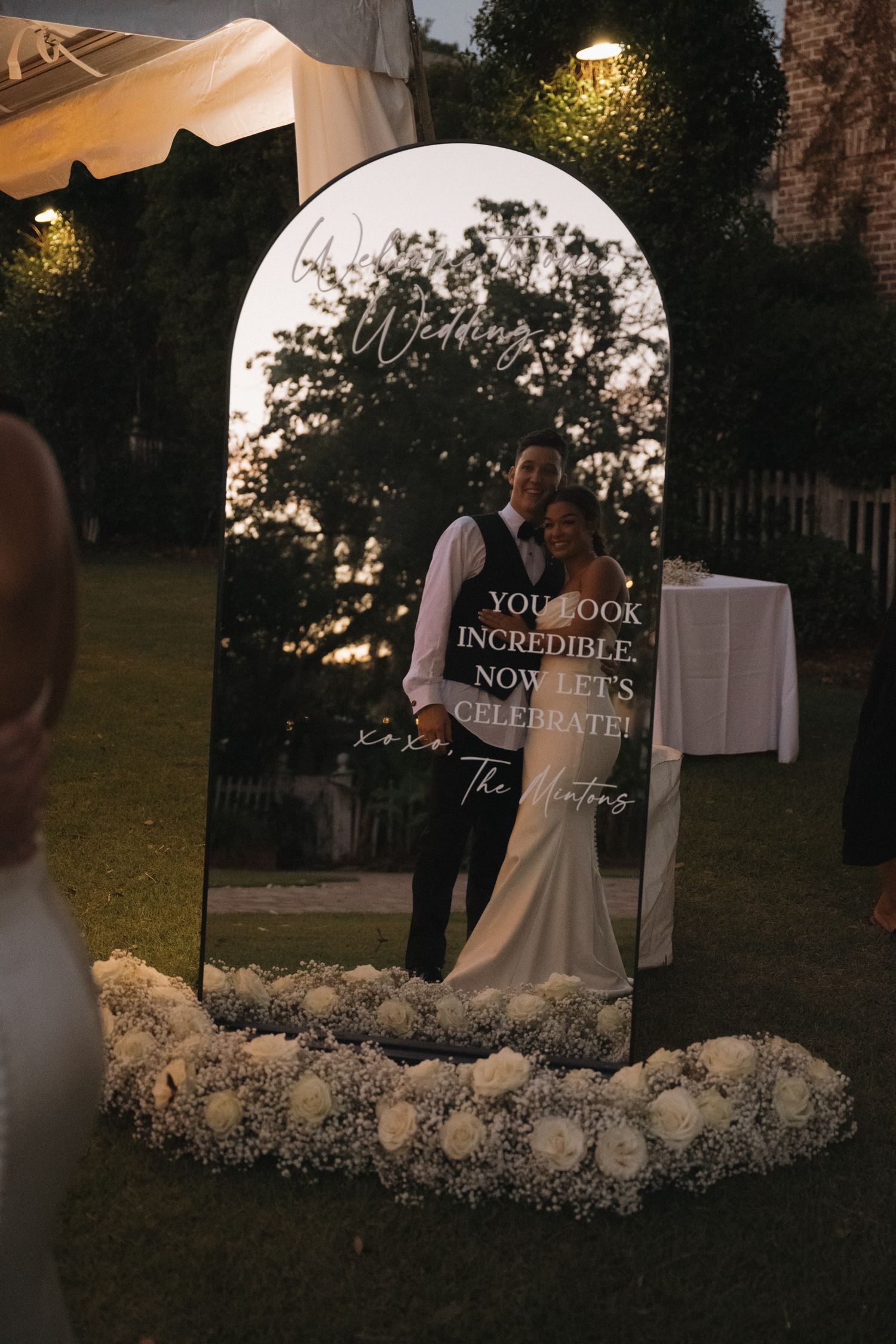 Wedding welcome mirror sign with couple photo and white floral arrangement at outdoor evening reception.