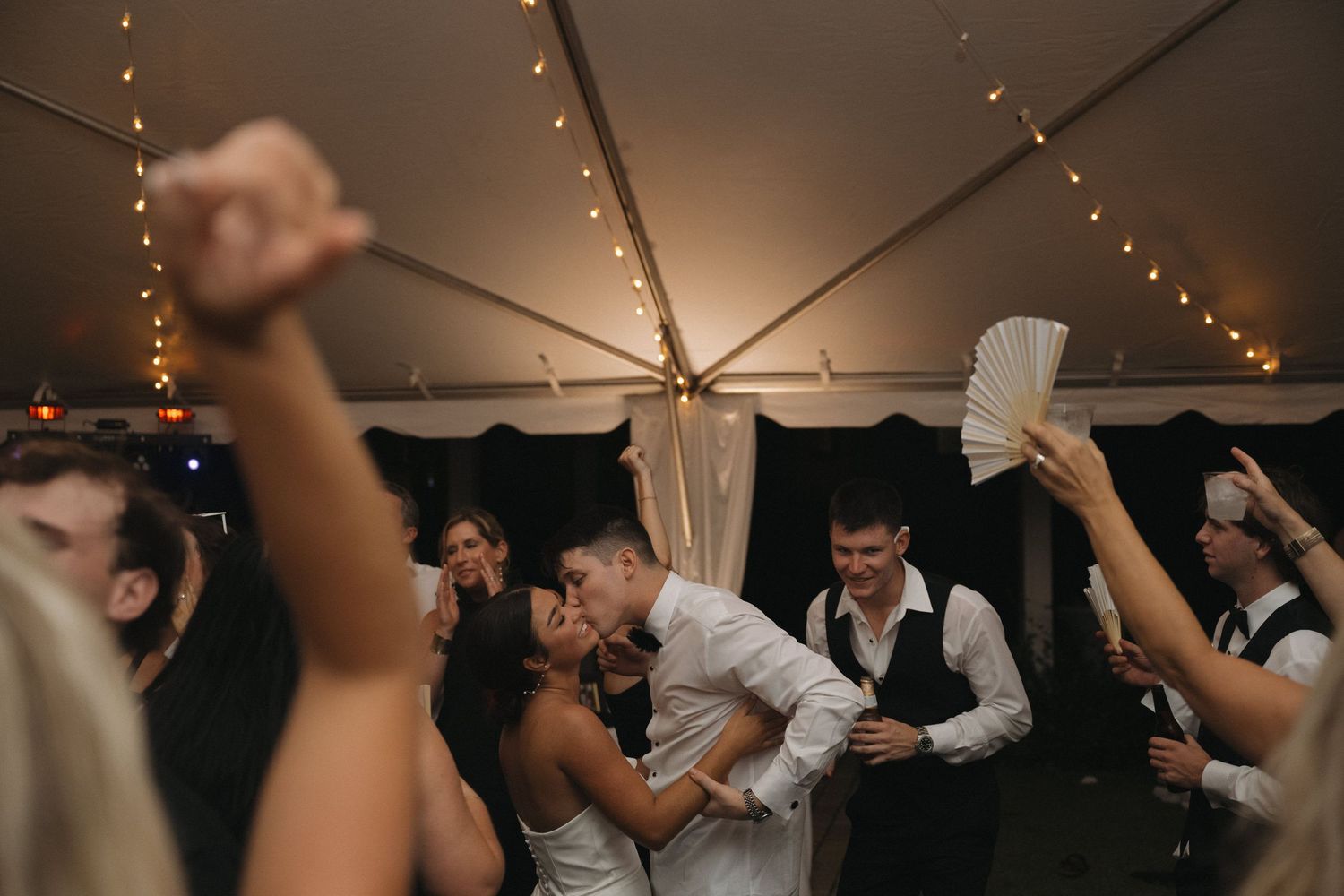 Bride and groom share a romantic kiss on the dance floor surrounded by cheering guests at a tented wedding reception.