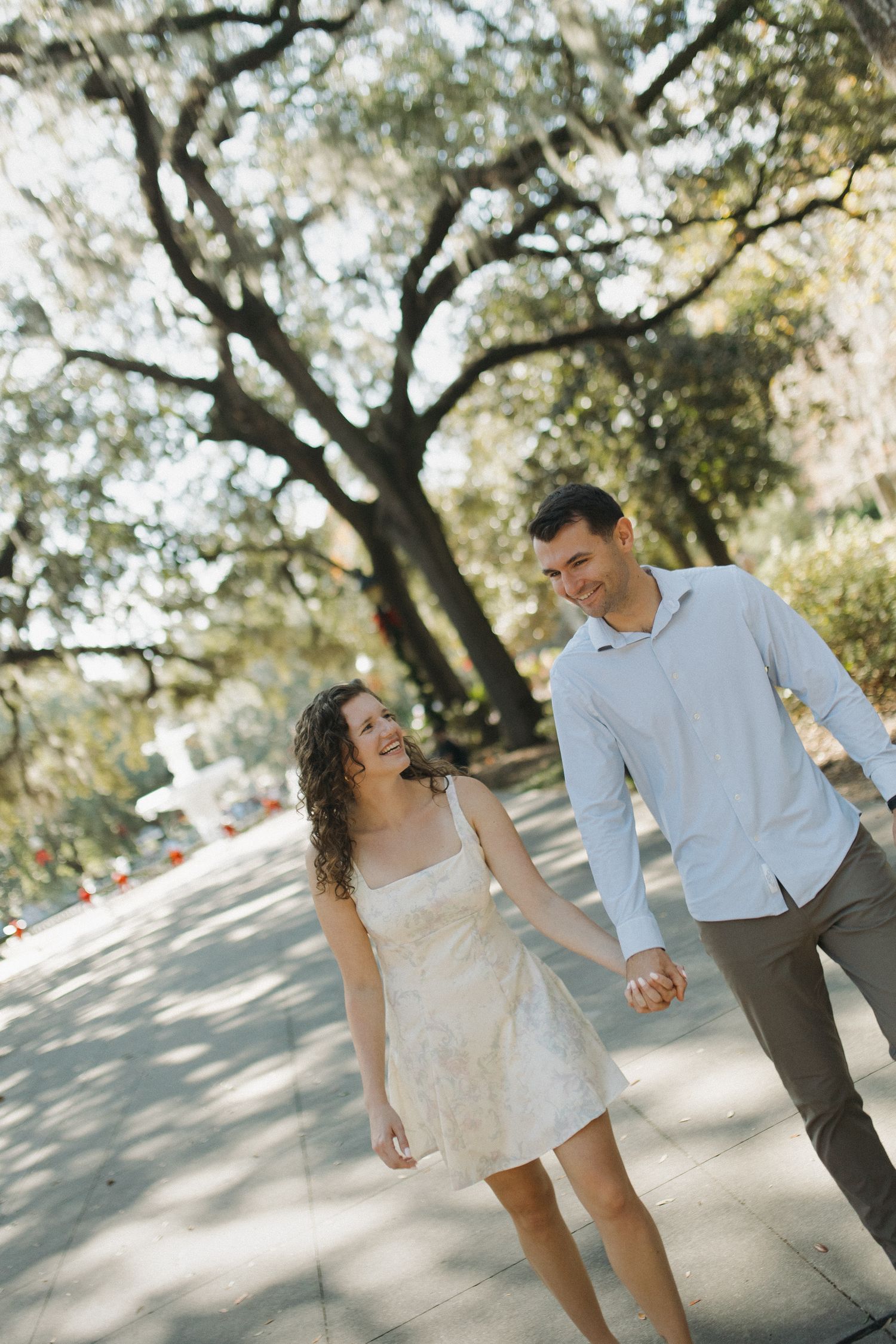 Happy couple holds hands walking along a sunlit path under large oak trees.