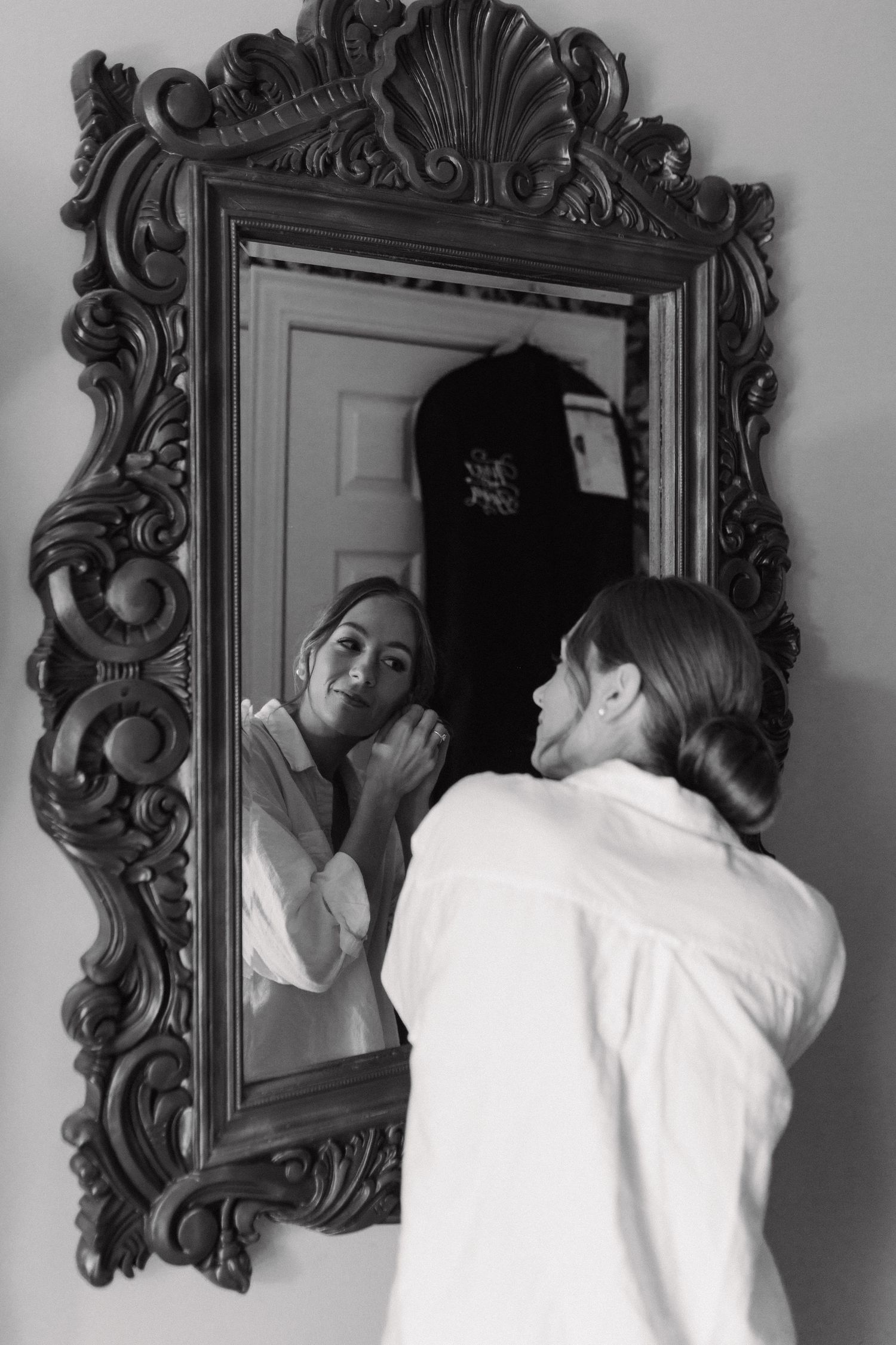 Black and white photo of a woman in a white robe adjusting her earrings in an ornate vintage mirror.