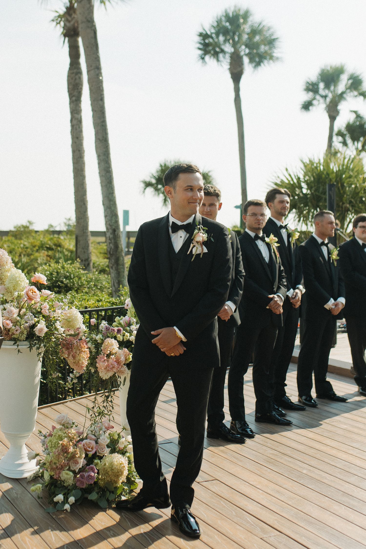 Groom in black tuxedo smiling at altar with groomsmen lined up behind, outdoor wedding ceremony with palm trees.