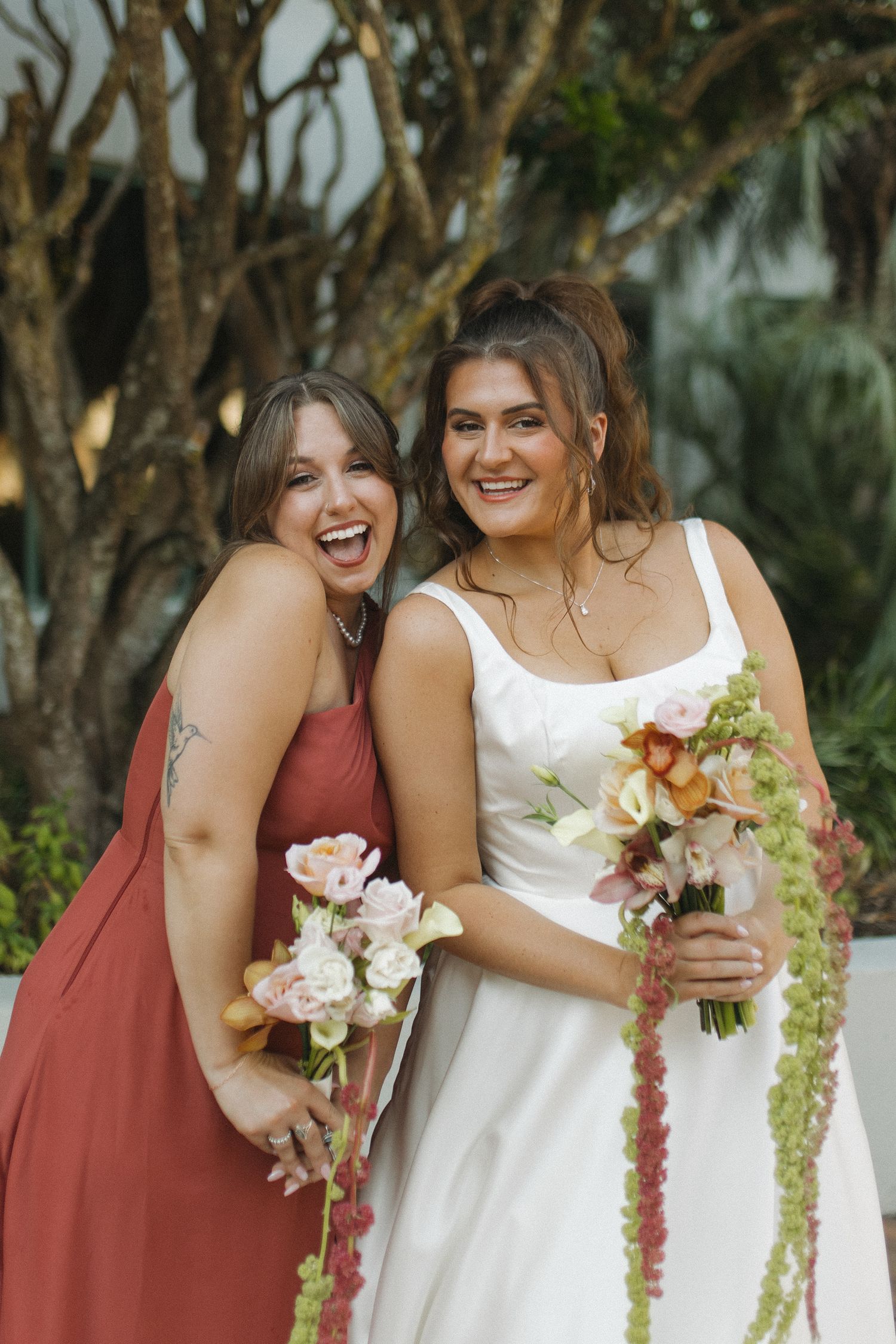 Smiling bride in white gown embraces bridesmaid in rust dress, both holding floral bouquets outdoors.