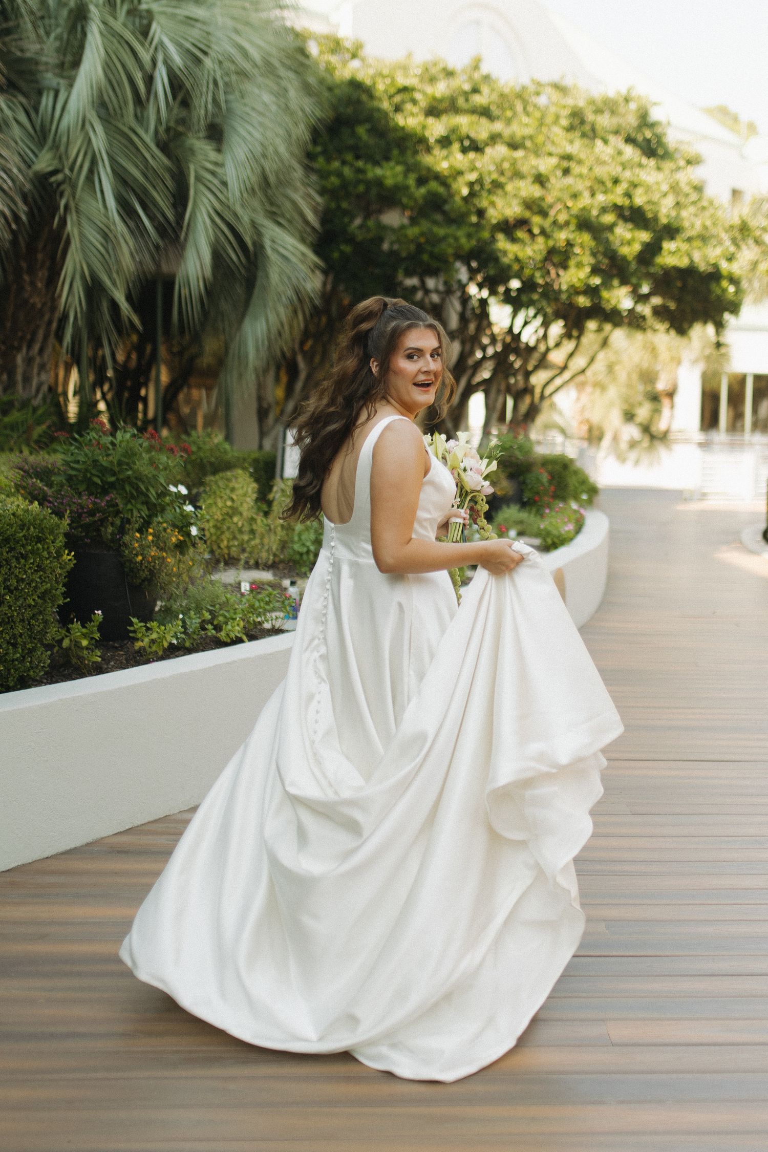 A joyful bride in a flowing white gown holds a bouquet while twirling on a wooden deck outdoors.