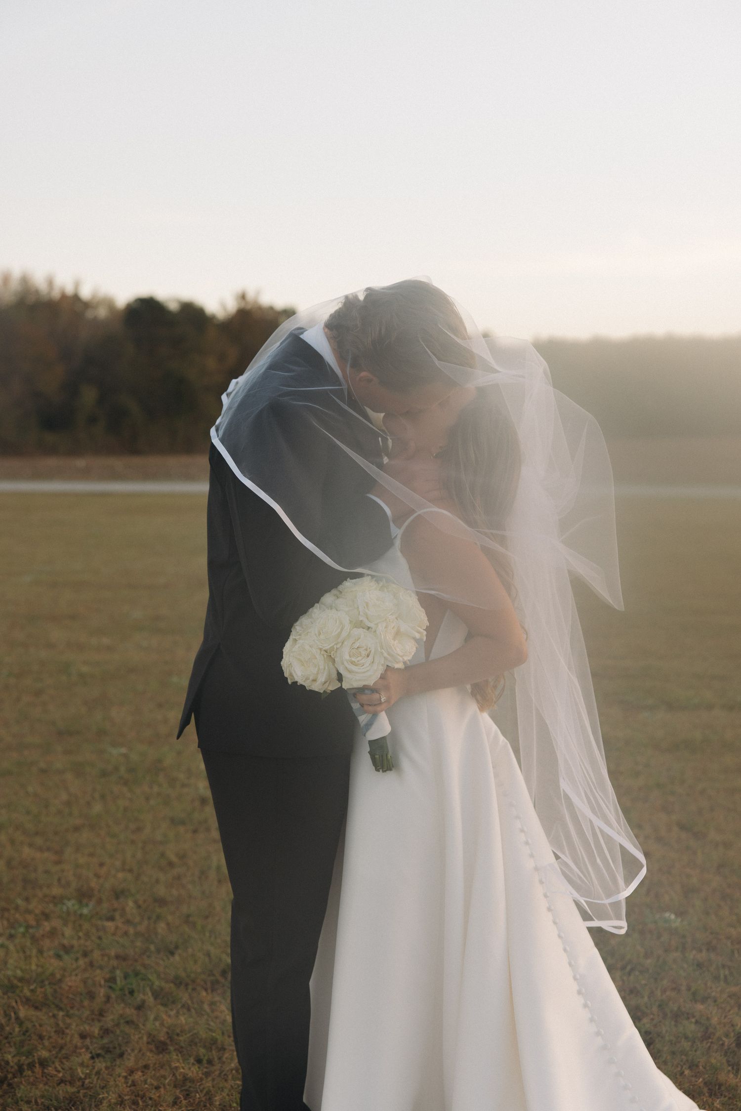Bride and groom sharing a kiss under a flowing white veil in an open field during golden hour.