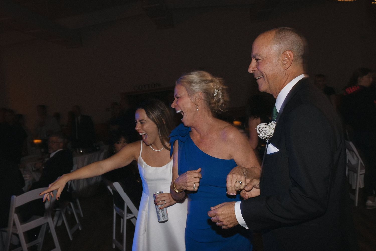 Three people dancing joyfully at a wedding reception, woman in blue dress linking arms with friends on dance floor.