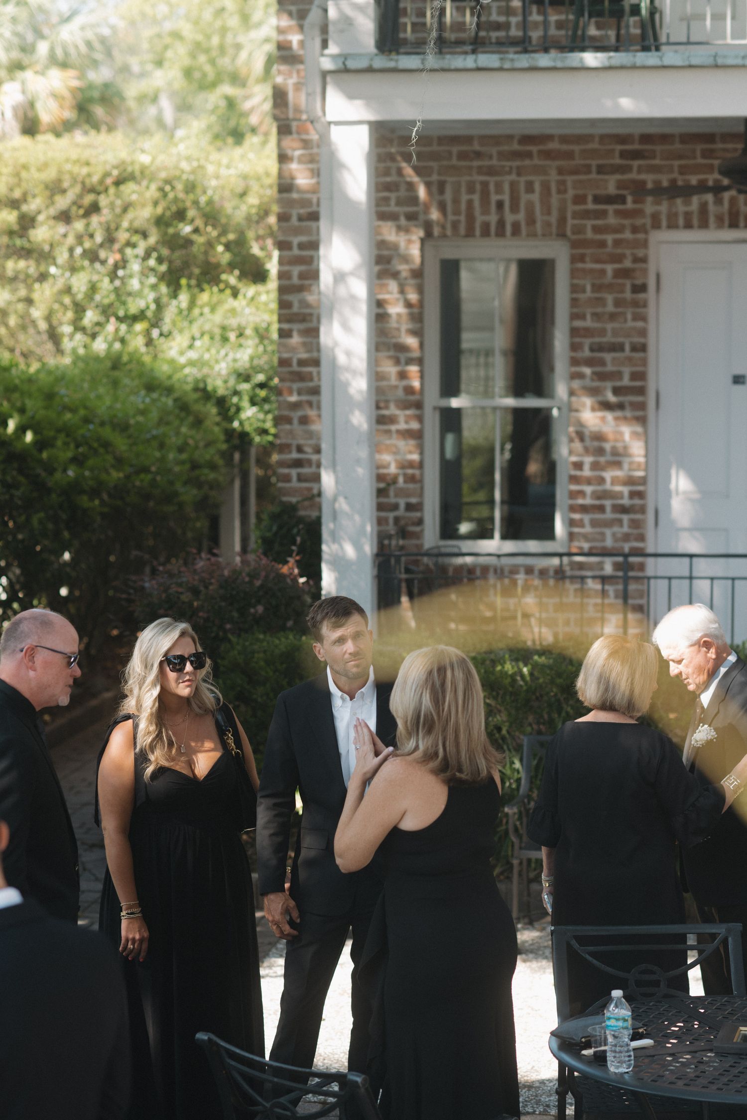 Wedding guests in black attire socializing outdoors near brick venue in sunlight.