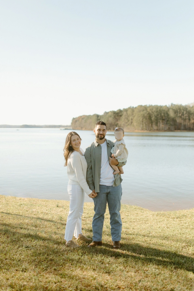 A smiling family of three poses by a serene lake, with parents holding their baby on a sunny day.