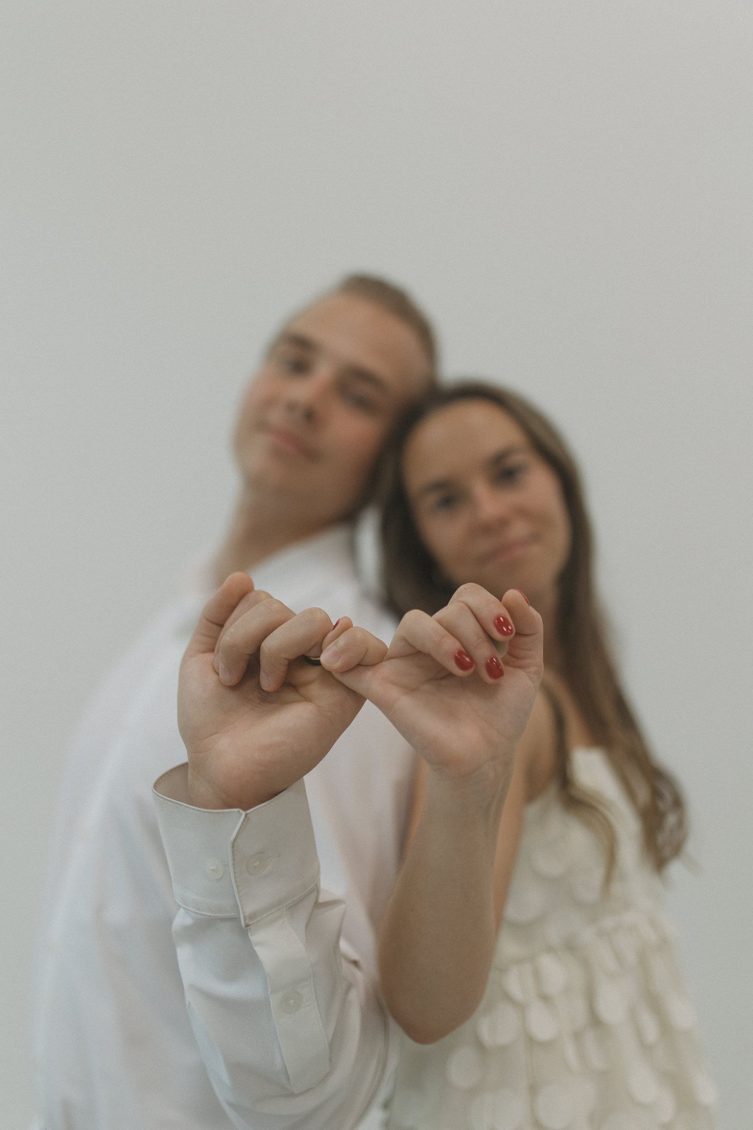 Couple holding pinkies together in foreground, wearing white outfits, smiling softly against white background.