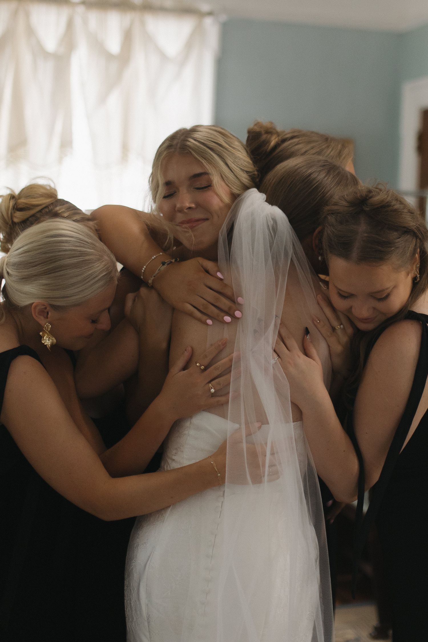 Bride in white gown and veil surrounded by bridesmaids in black dresses sharing a group hug.