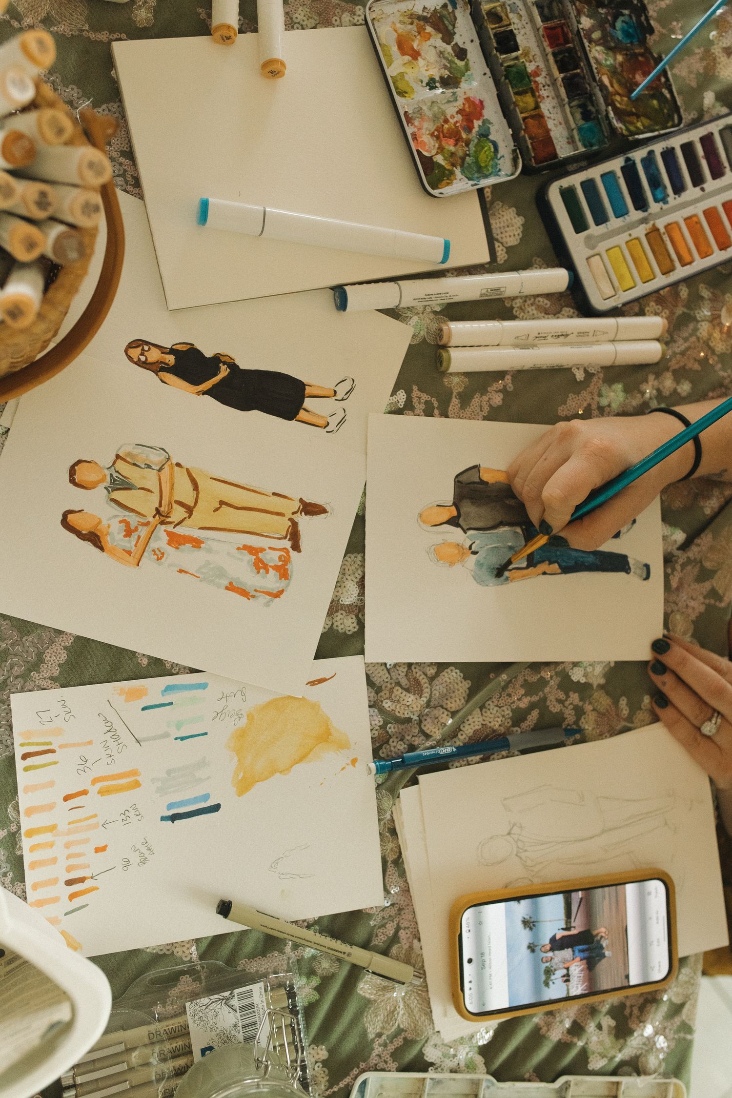 Fashion designer sketching clothing illustrations with watercolors and markers on a cluttered desk.