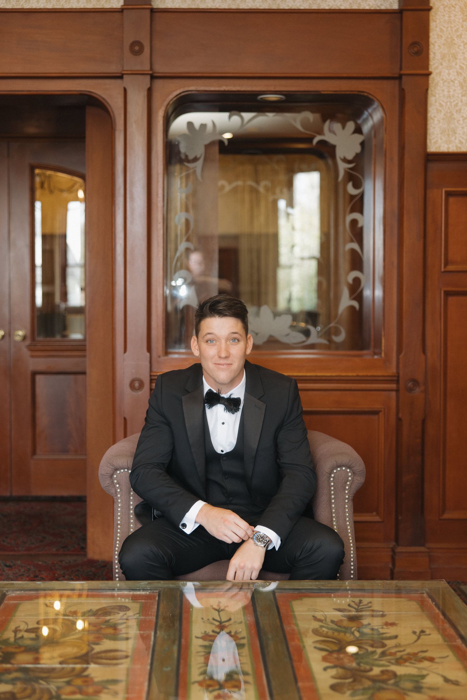 Dapper groom in black tuxedo with bow tie seated confidently in ornate wooden chair in classic interior setting.