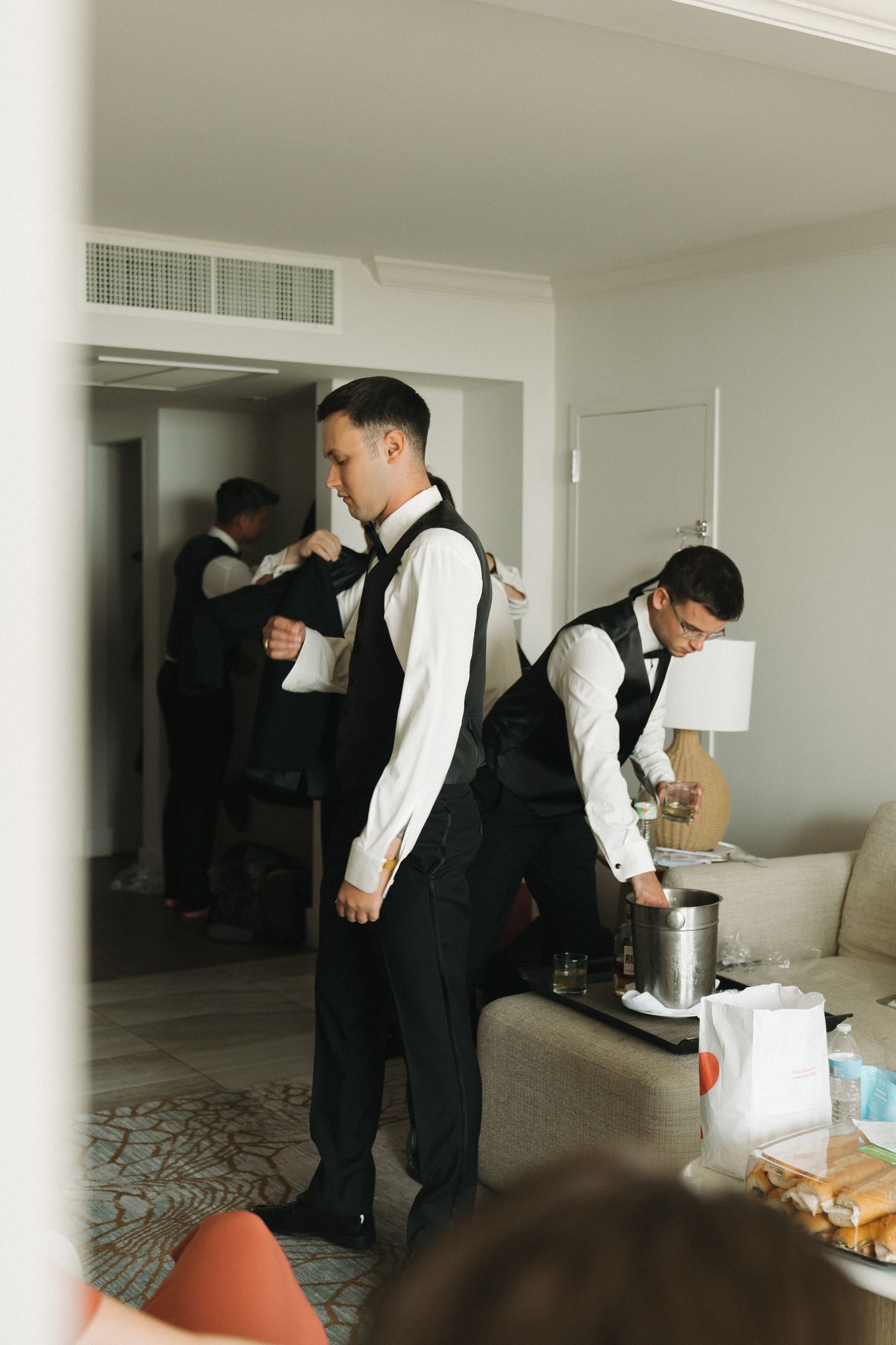 Groomsmen in black vests and white shirts preparing for a wedding ceremony in a bright hotel room.
