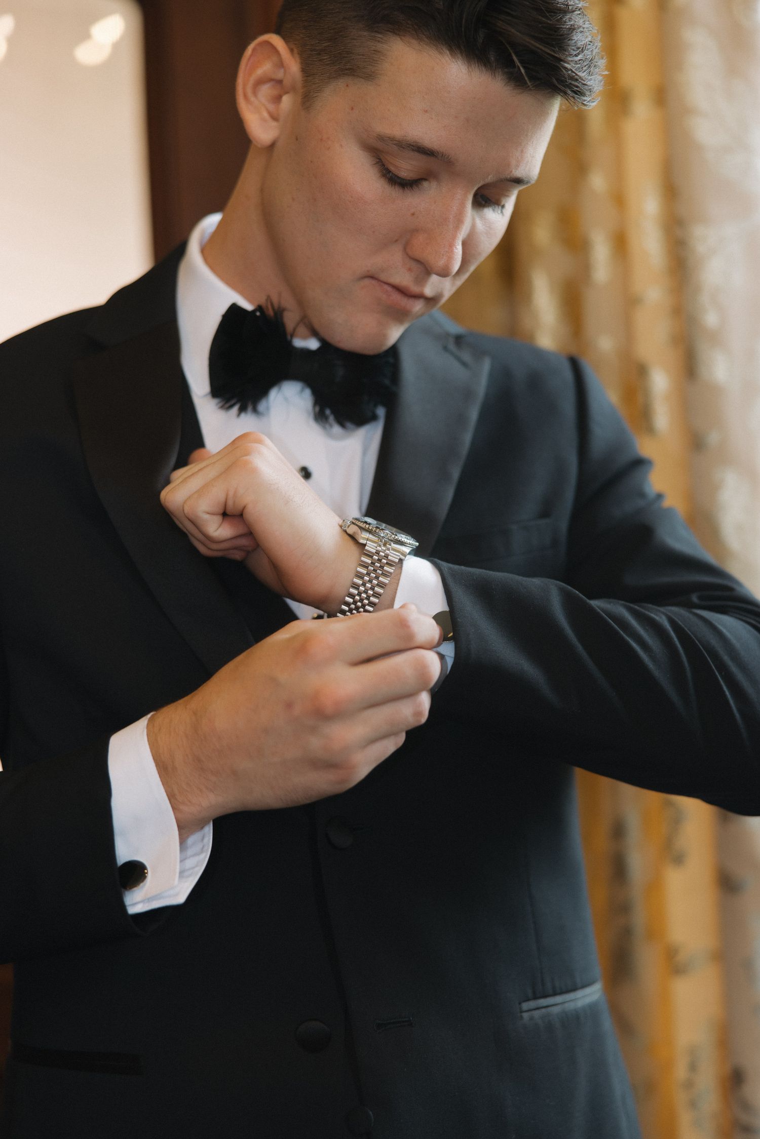 Groom in black tuxedo and bow tie adjusting his silver watch cufflinks while getting ready for wedding.