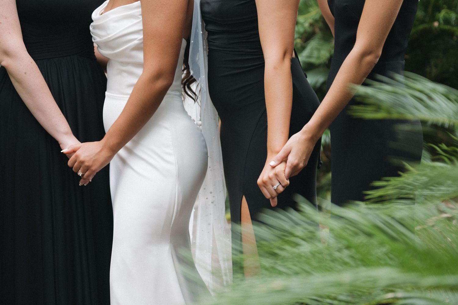 Bride in white gown holds hands with bridesmaids in black dresses, surrounded by lush green foliage.