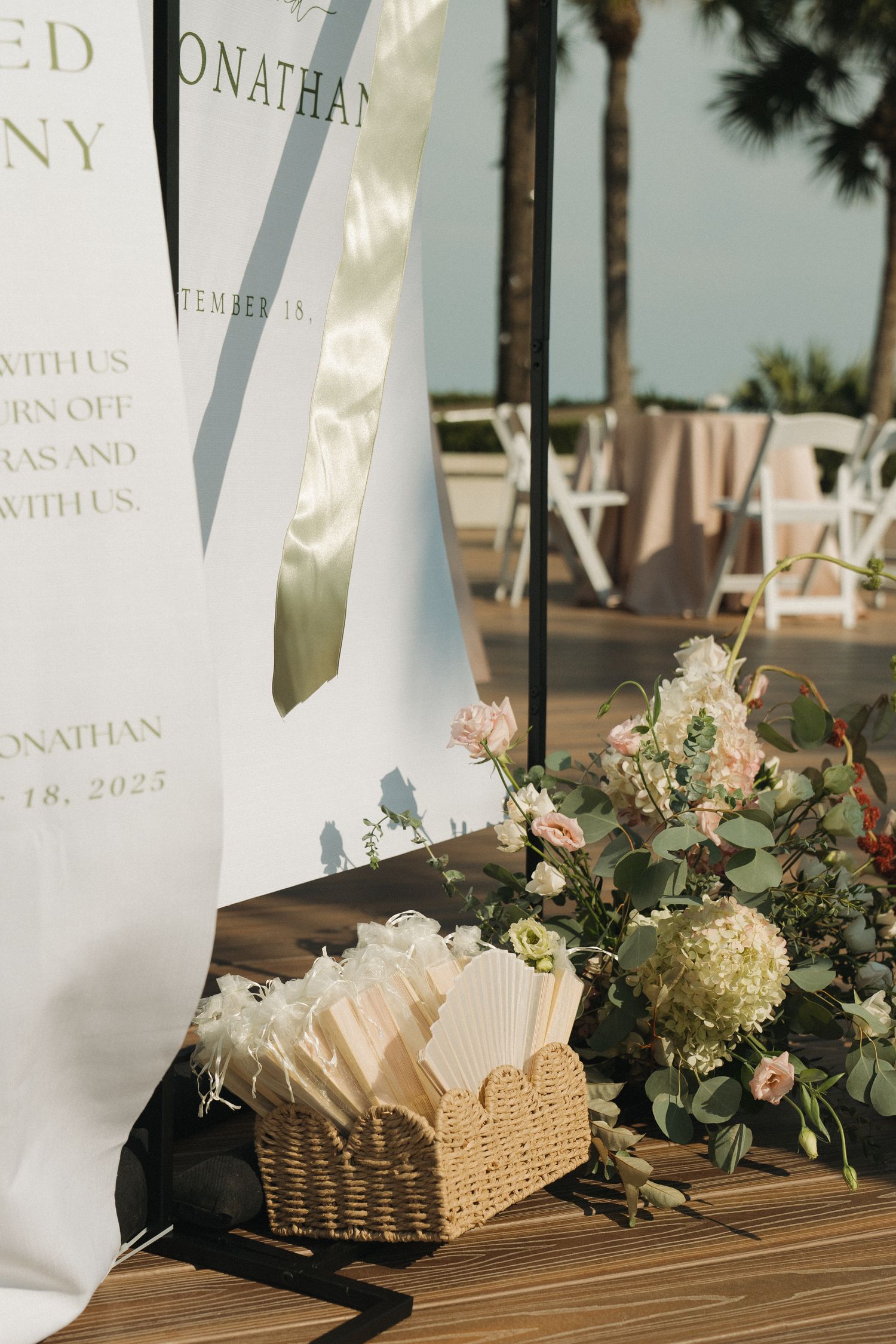 Outdoor wedding ceremony setup with white fans in a wicker basket, floral arrangements, and a personalized welcome sign.