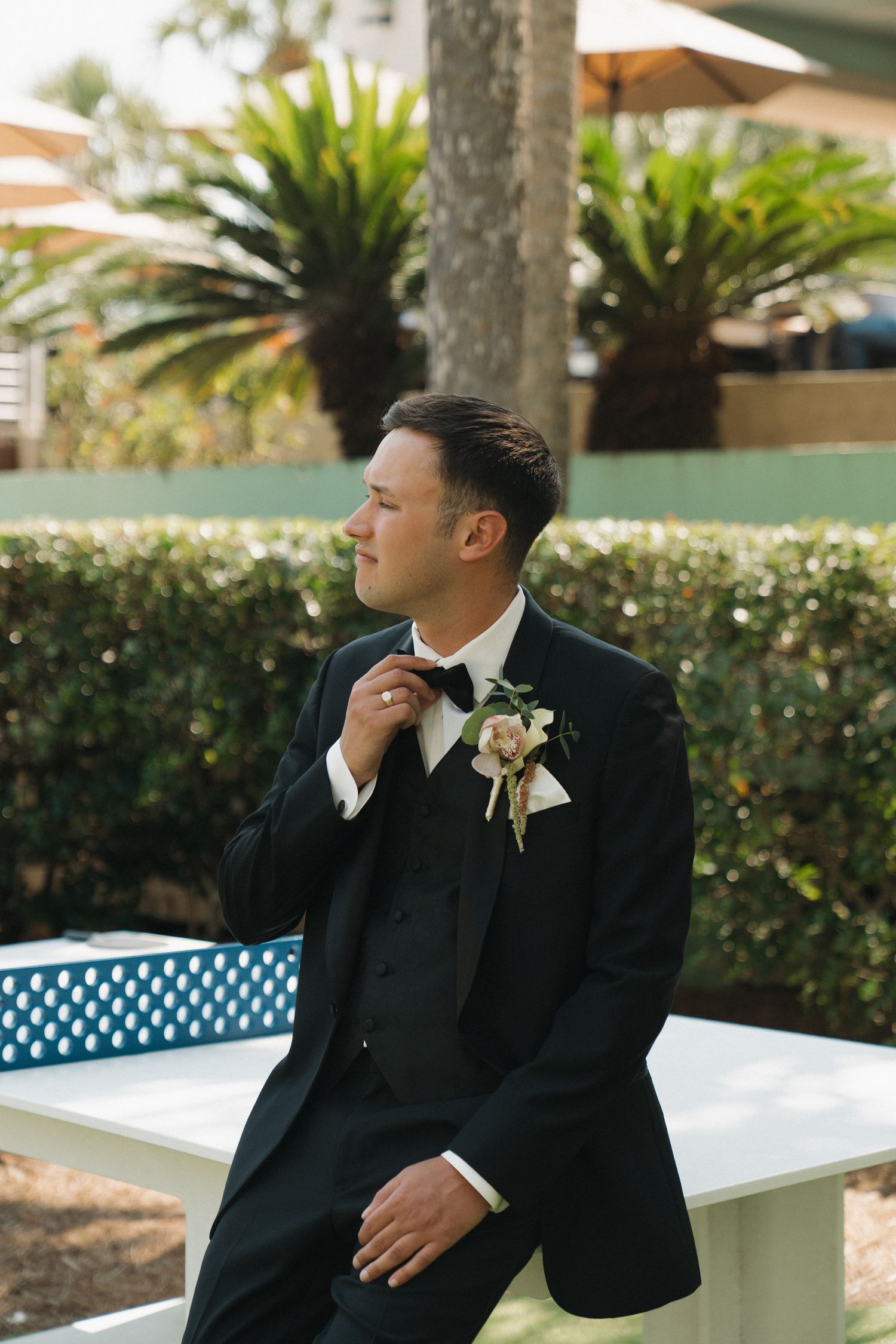 Groom in black tuxedo adjusting bow tie while seated outdoors with tropical greenery background.