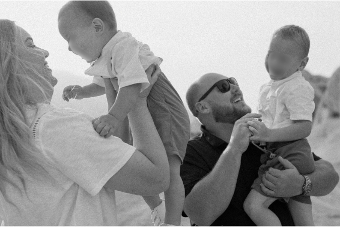 Black and white photo of a happy family with two babies on a beach, parents lifting and holding infants joyfully.