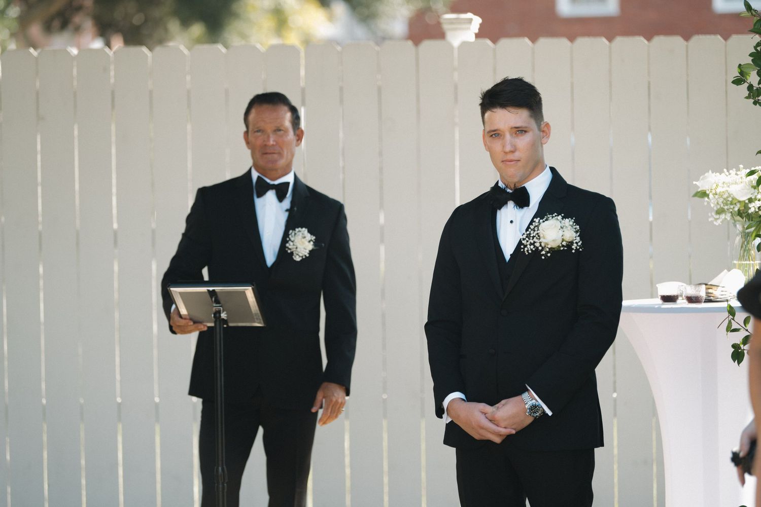 Groom in black tuxedo stands at outdoor wedding altar with officiant beside white fence backdrop.
