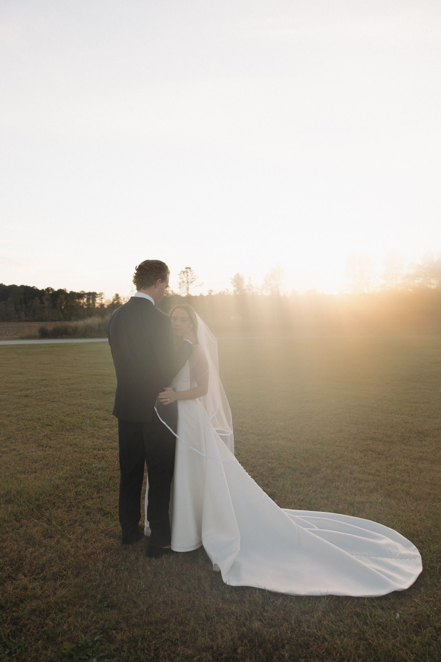 Bride and groom silhouetted against a golden sunset in an open field, bride wearing a long cathedral veil.