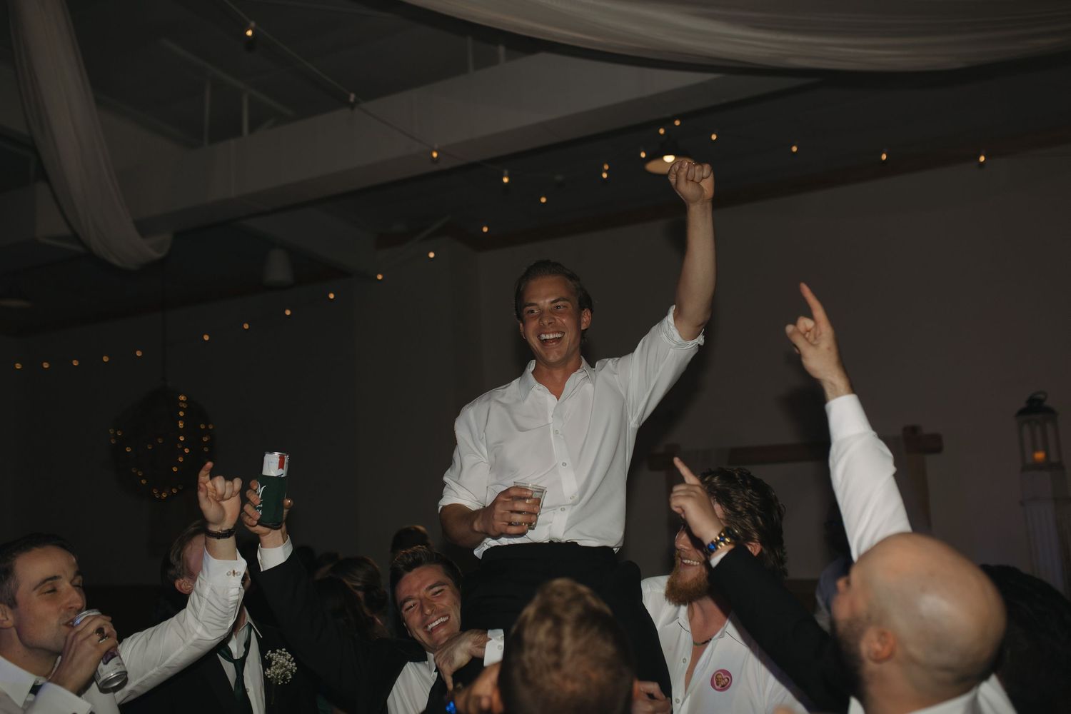 Groom lifted by groomsmen celebrating at wedding reception party indoors.