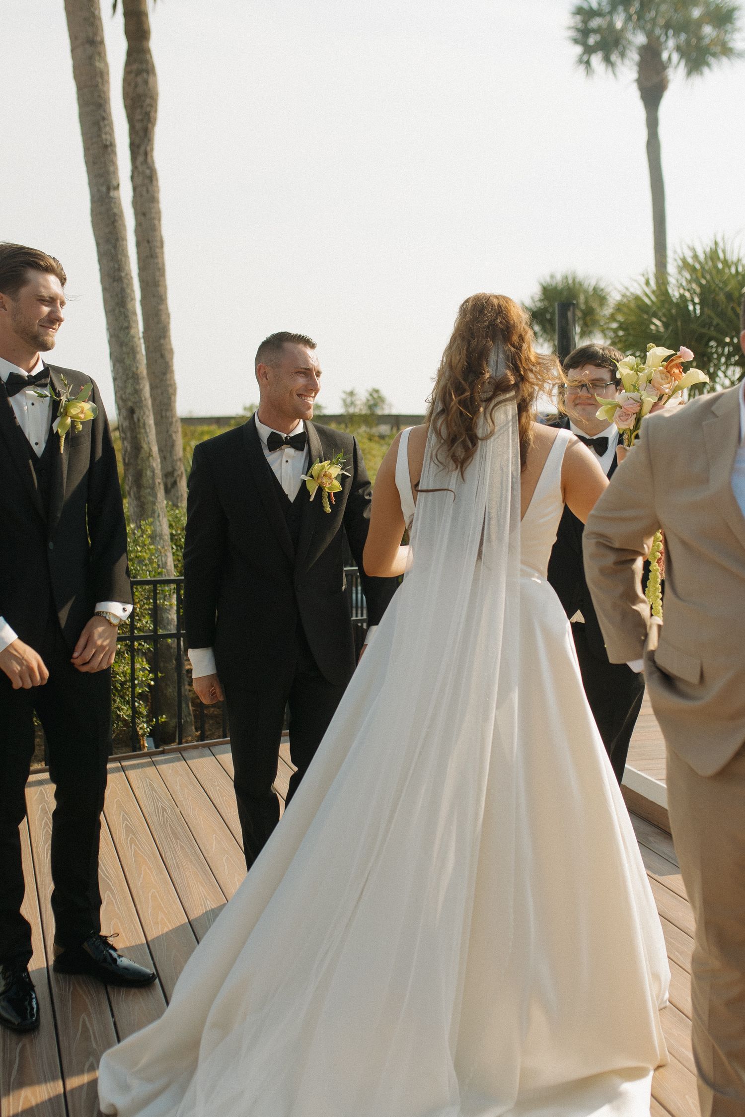 Bride in white gown with veil faces smiling groom in black tuxedo during outdoor wedding ceremony on wooden deck.