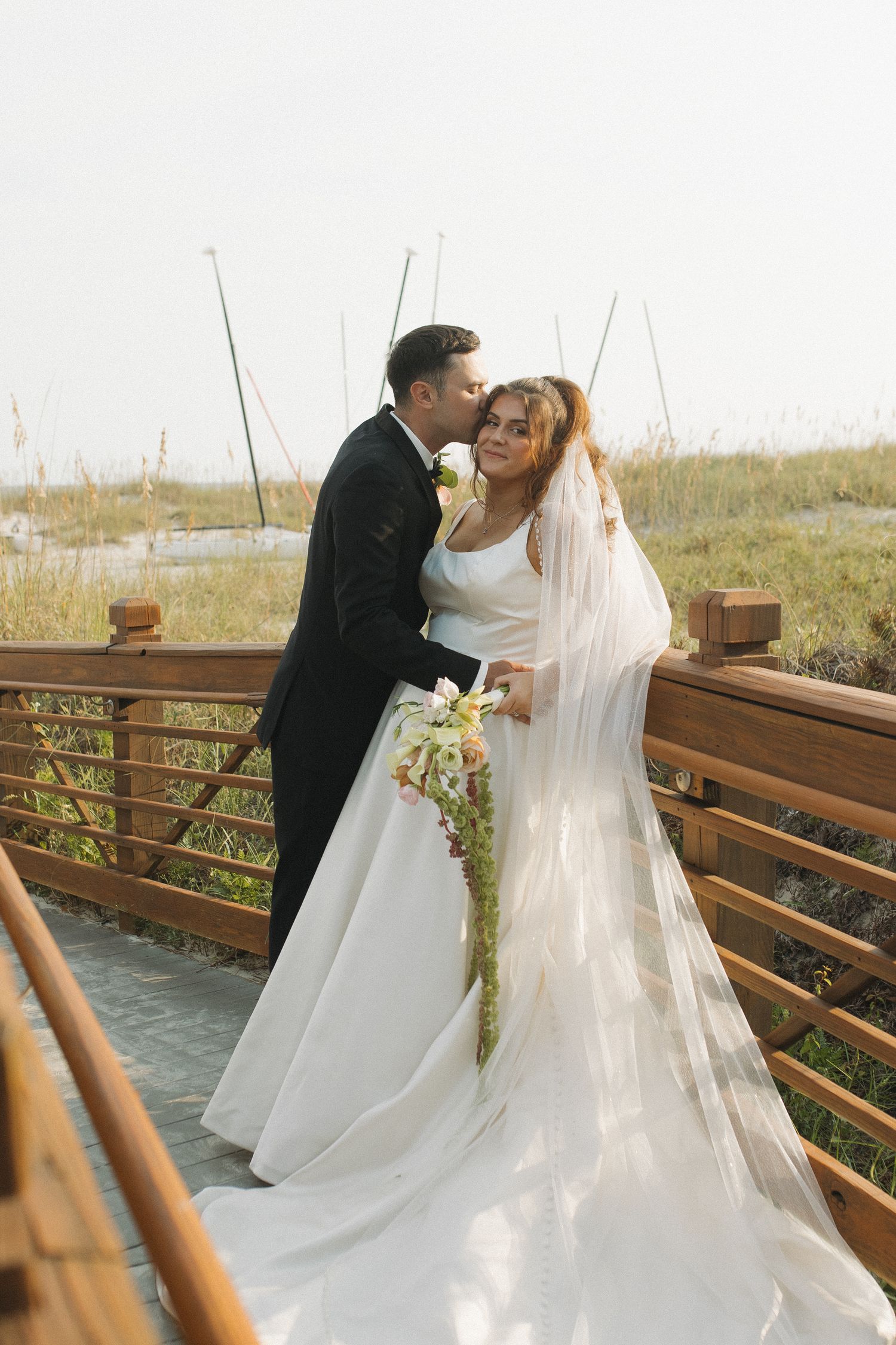 Bride and groom share a romantic kiss on a wooden boardwalk, bride holding a cascading bouquet in a white gown and veil.