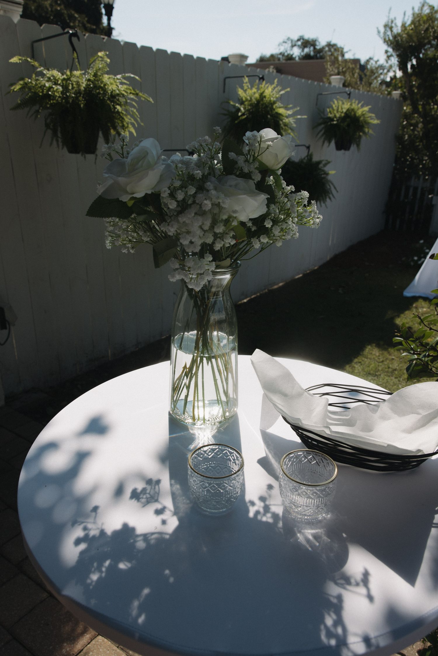Outdoor reception table with white linen and glass vase of white roses and greenery in sunny backyard garden.