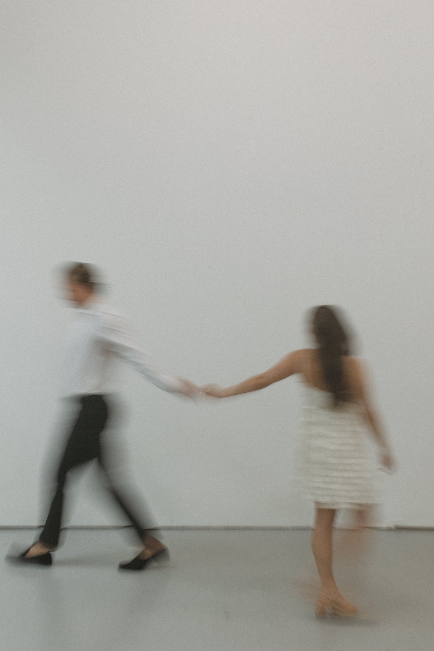 Blurred motion photo of couple holding hands walking in minimalist white studio space.