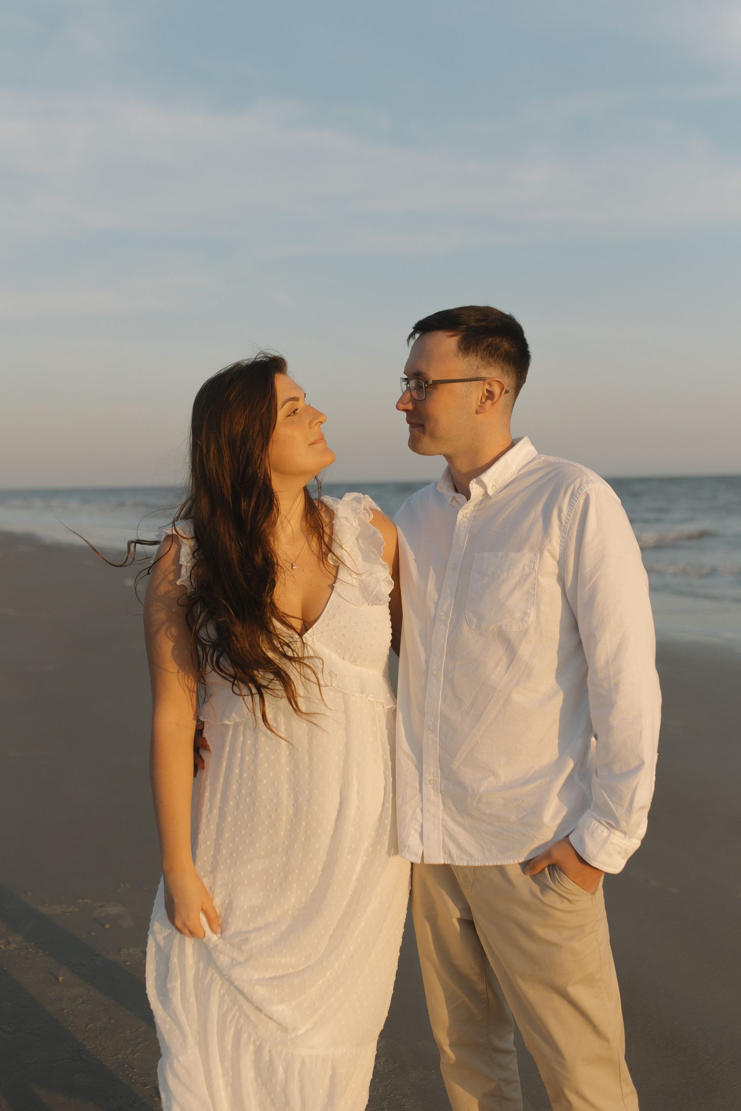 Couple in white outfits sharing a romantic moment on the beach at golden hour.