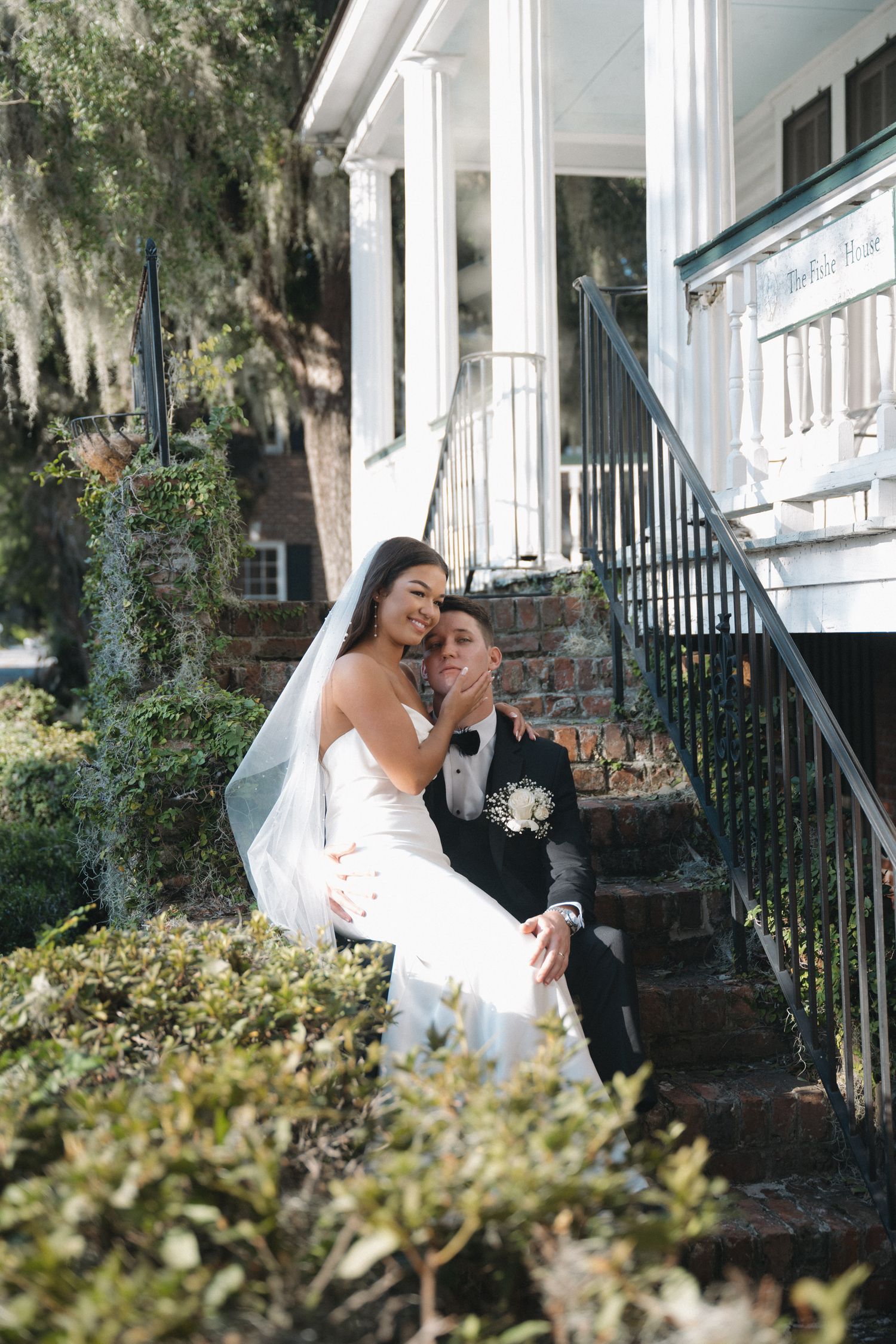 Bride and groom sitting on historic home steps, bride whispering to groom under Spanish moss trees.
