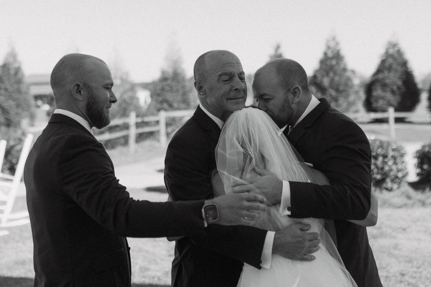 Black and white wedding photo of bride embracing groom while two men in suits stand nearby outdoors.