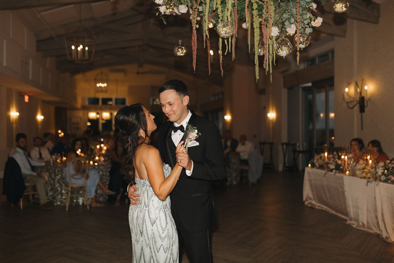 Bride and groom share their first dance under floral chandelier at elegant candlelit wedding reception.
