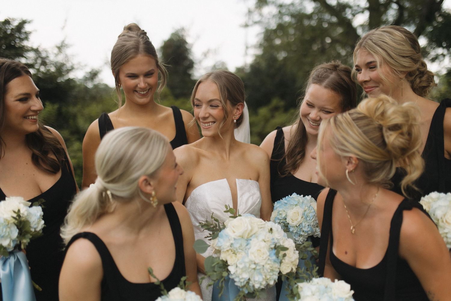 Bride surrounded by bridesmaids in black dresses holding white and blue bouquets.