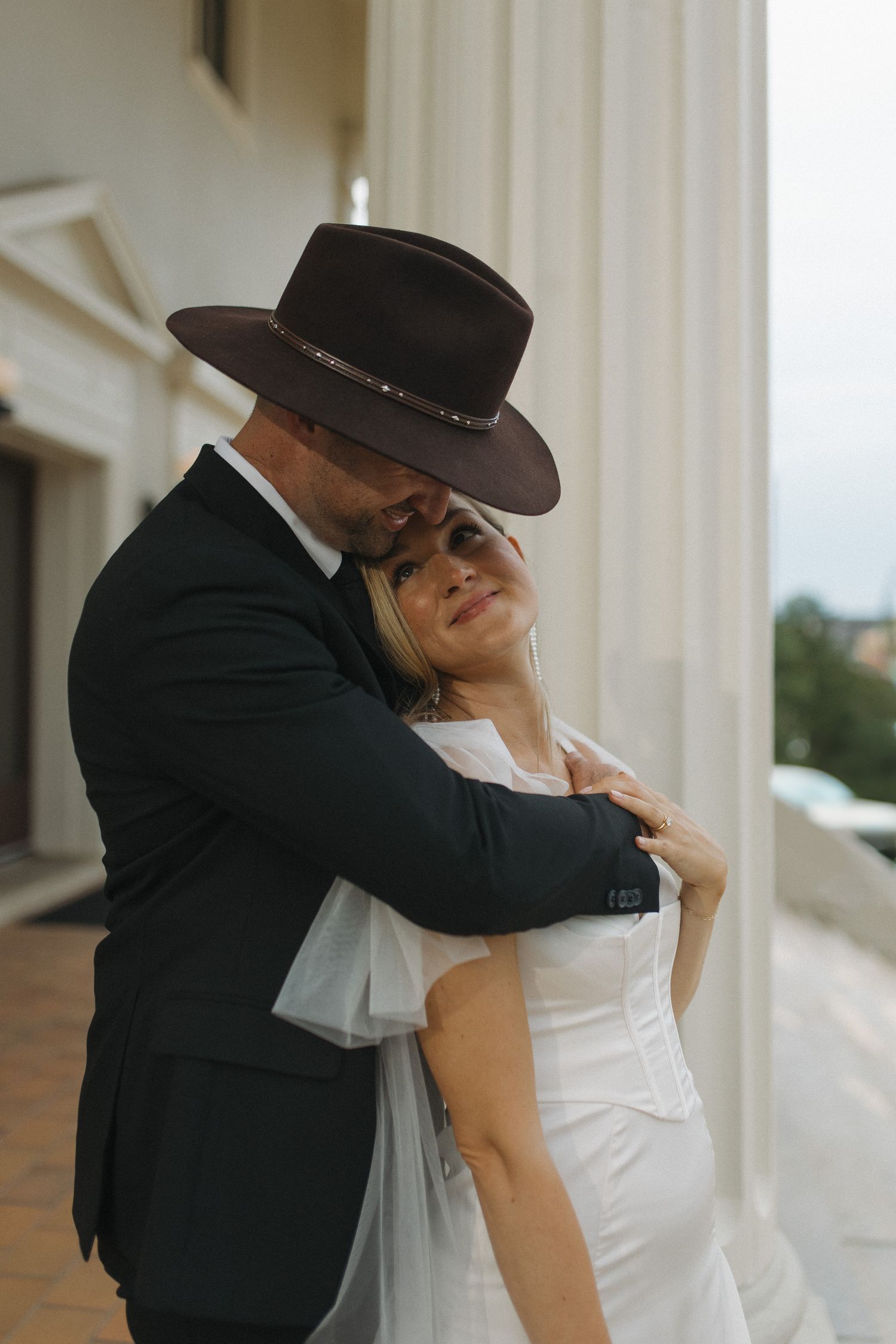 Bride and groom embrace near white columns, groom in dark suit and brown wide-brim hat kissing brides temple.