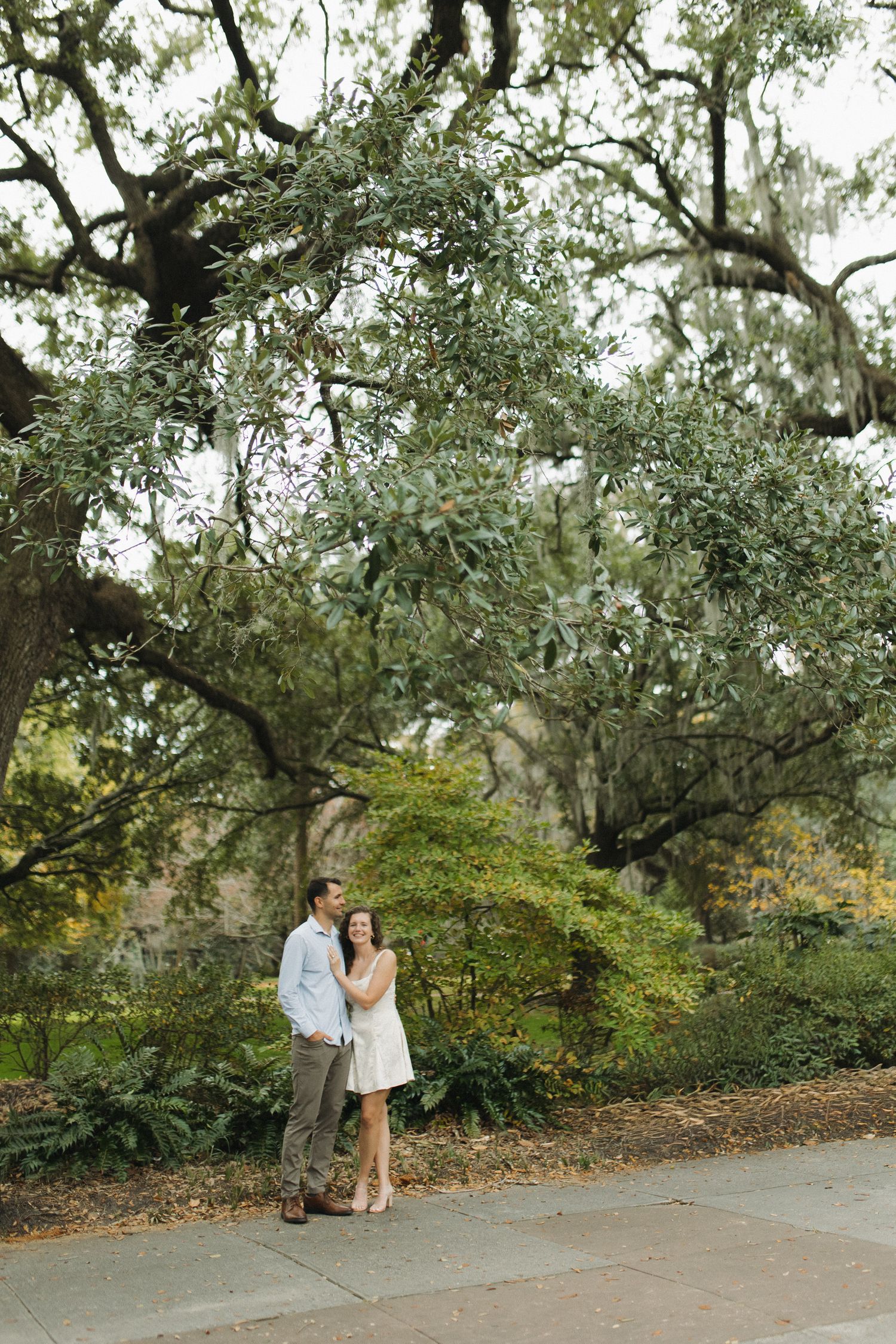 Couple embraces under sprawling moss-covered oak trees in a lush garden setting.