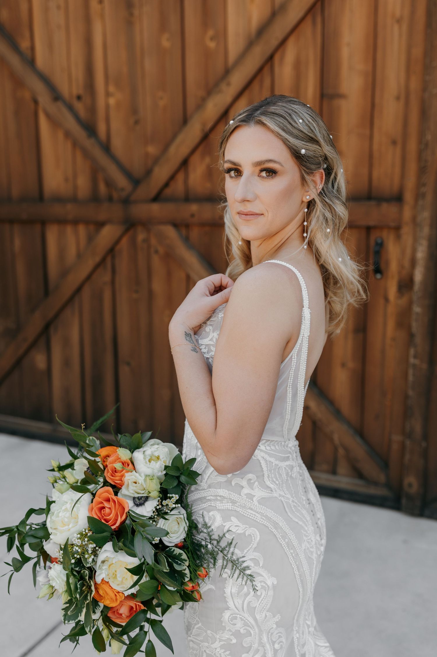 Elegant bride in lace gown holds colorful orange and white bouquet before rustic barn doors.