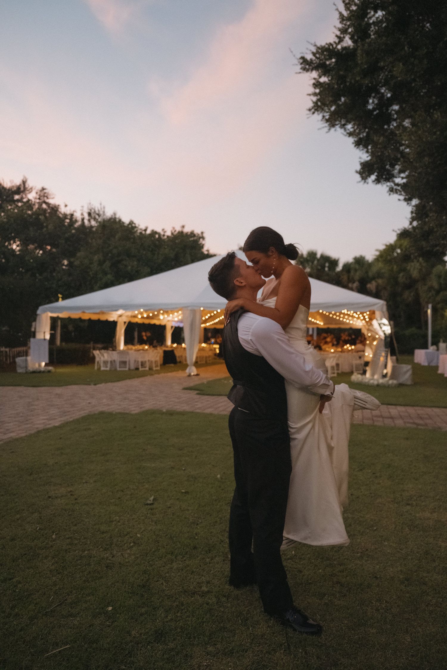 Groom lifts bride in white gown for a romantic kiss at dusk with a softly lit reception tent in the background.