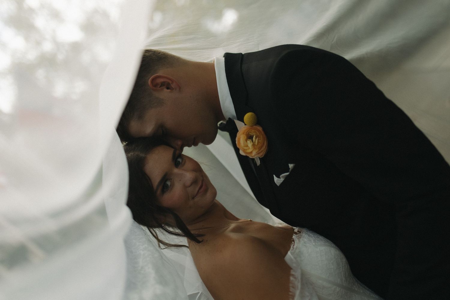 A bride and groom share an intimate forehead-to-forehead moment under a flowing white veil, groom wearing a yellow boutonniere.